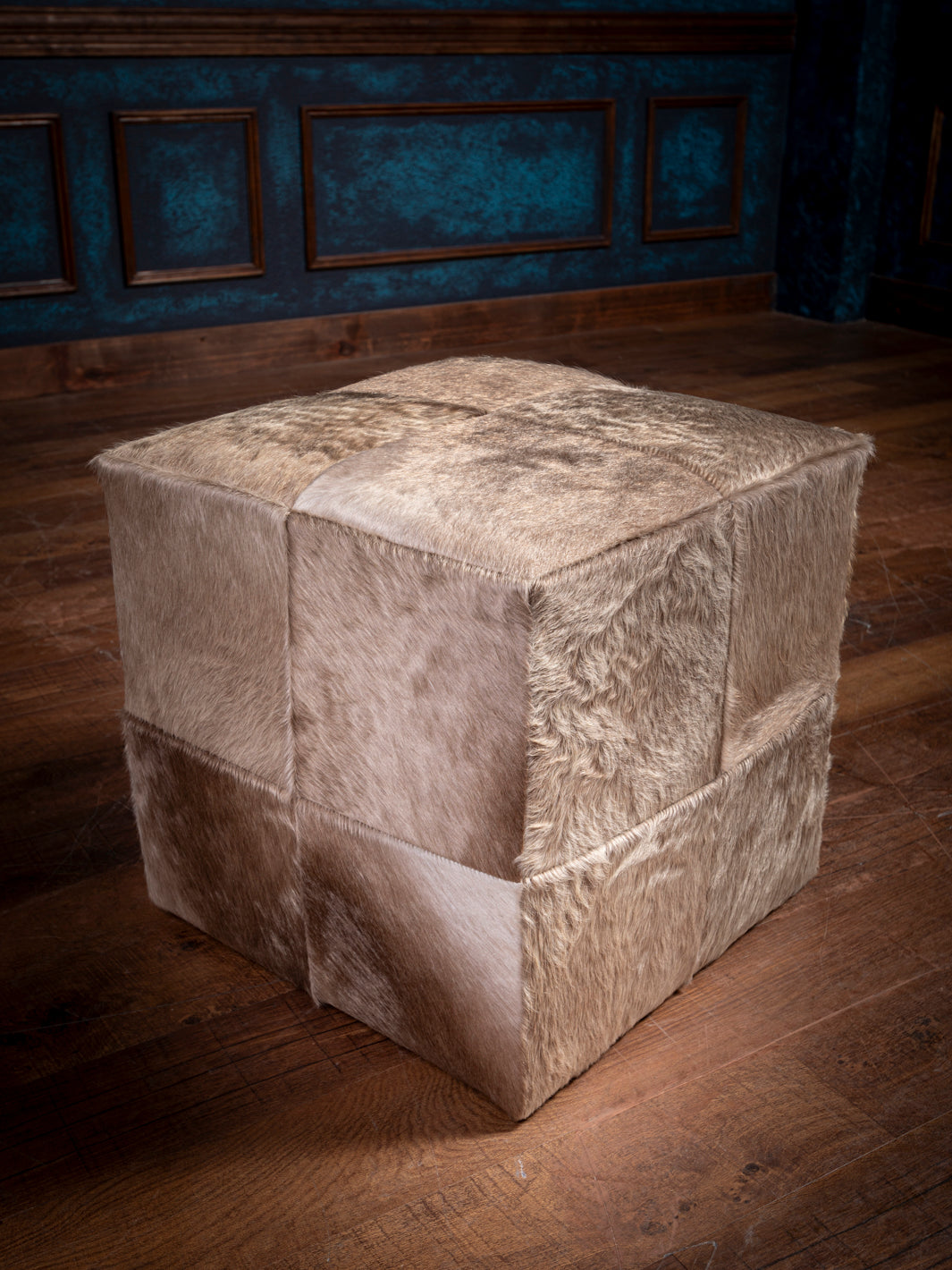 Beige hair on hide cube ottoman on a wooden floor, with teal paneled walls and brown trim in the background.