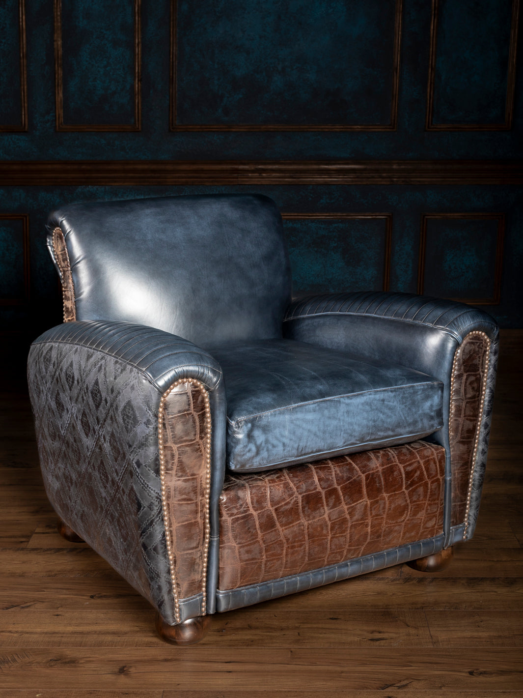 A blue leather cigar chair with embossed details and croc-embossed accents, featuring a structured silhouette and aged nailhead trim, set against a dark paneled wall on a wooden floor.