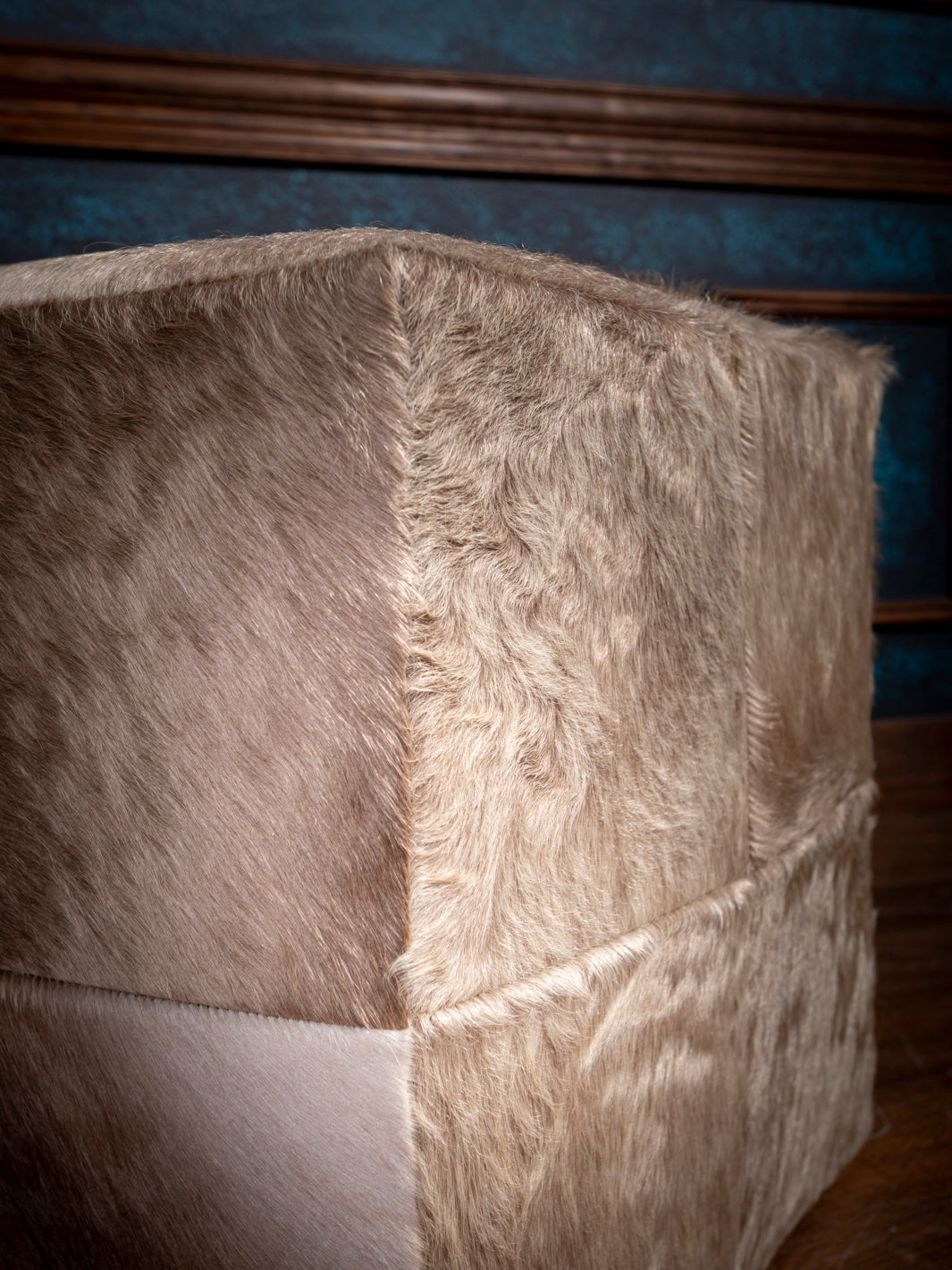 Close-up of a beige faux fur cube ottoman against teal paneled walls with brown trim.