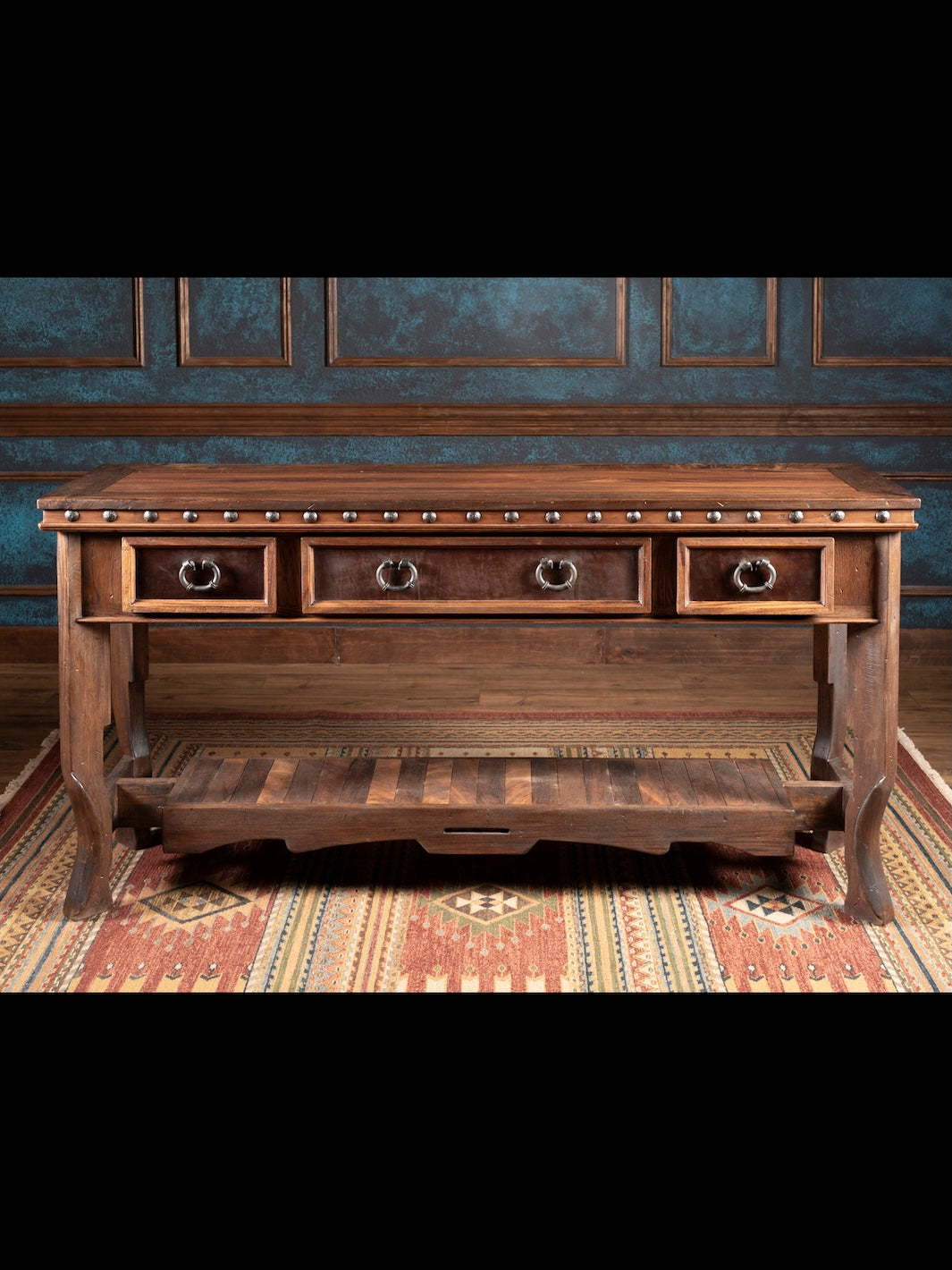 Wooden console table with three drawers in a room with patterned wallpaper and rug.
