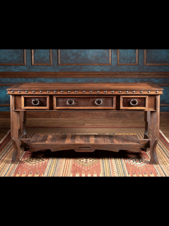 Wooden console table with three drawers in a room with patterned wallpaper and rug.