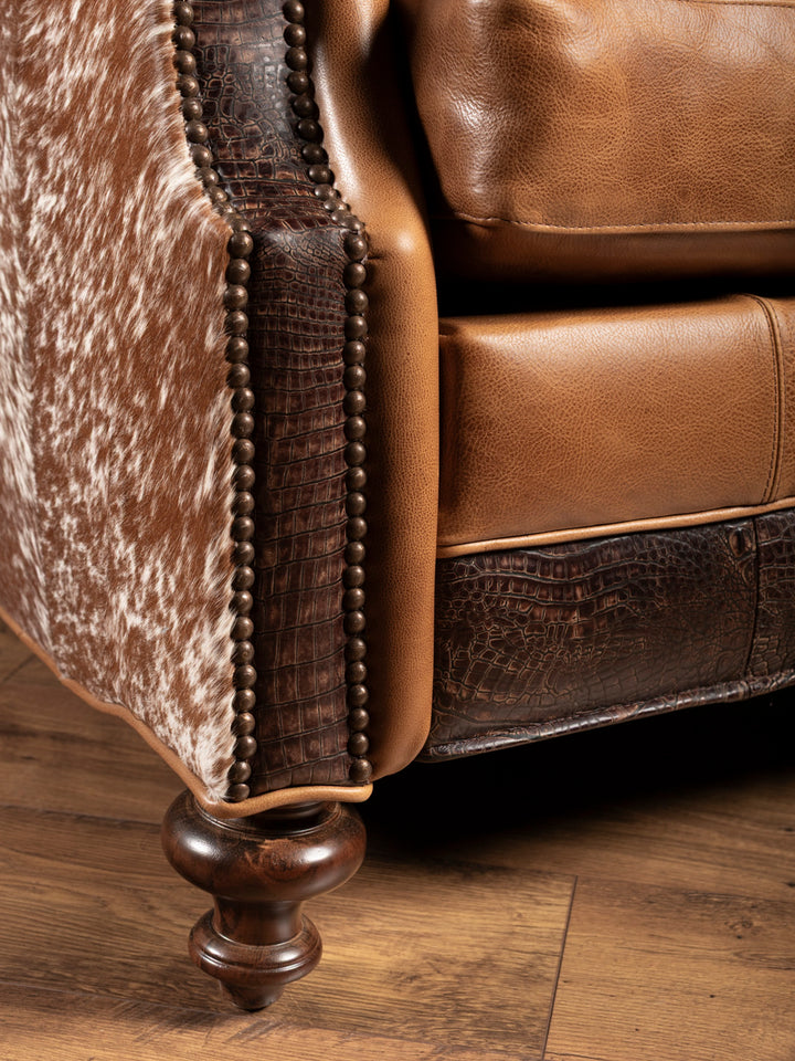 Close-up of chair base showcasing turned wood leg, crocodile-embossed leather, saddle leather cushion, and brindle cowhide side panel.