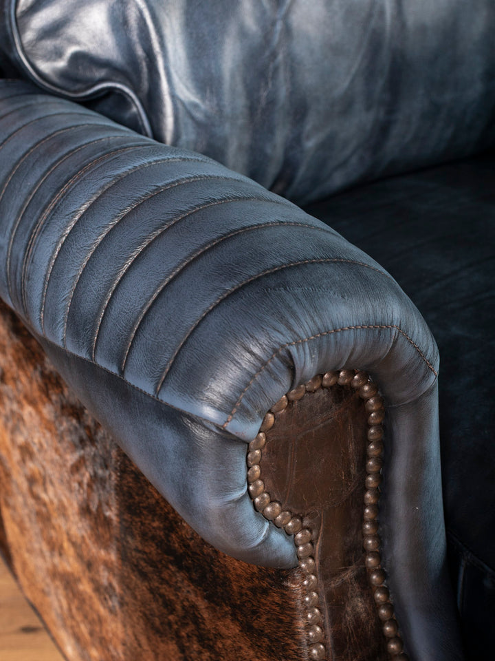 Blue leather recliner arm with boot stitch quilting, cowhide panel, and antique brass nailhead trim craftsmanship close-up