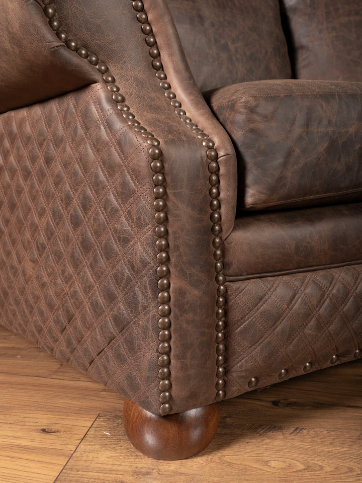 Close up angle of the sofa showcasing brown leather inlay, nailheads, and round bun feet 