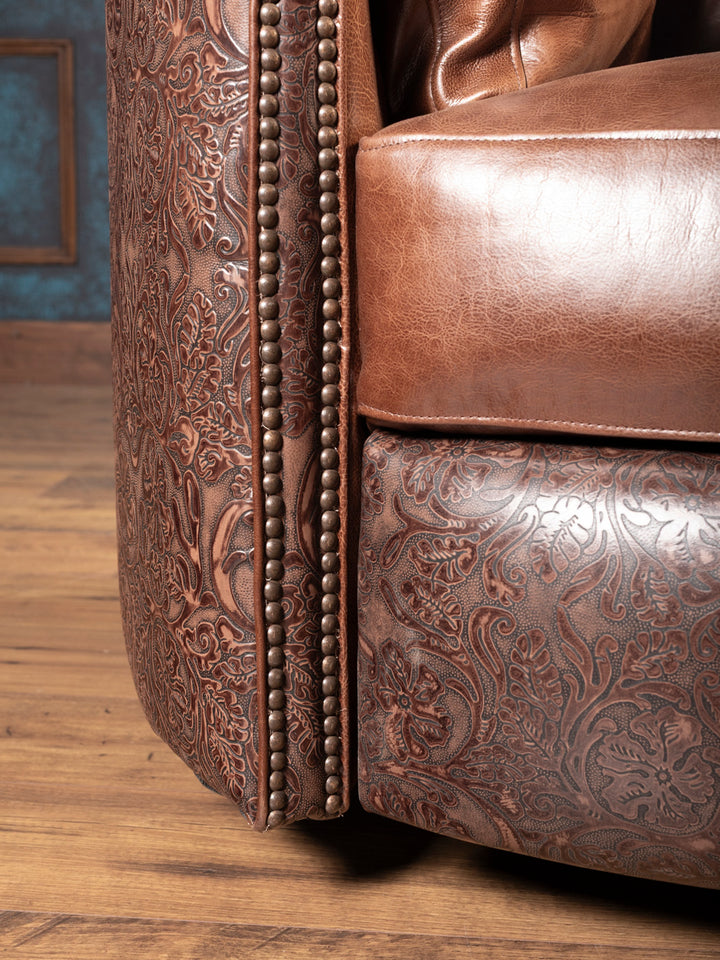 Close-up of arm detail showing tooled leather floral embossing, nailhead trim, and saddle leather seat cushion.