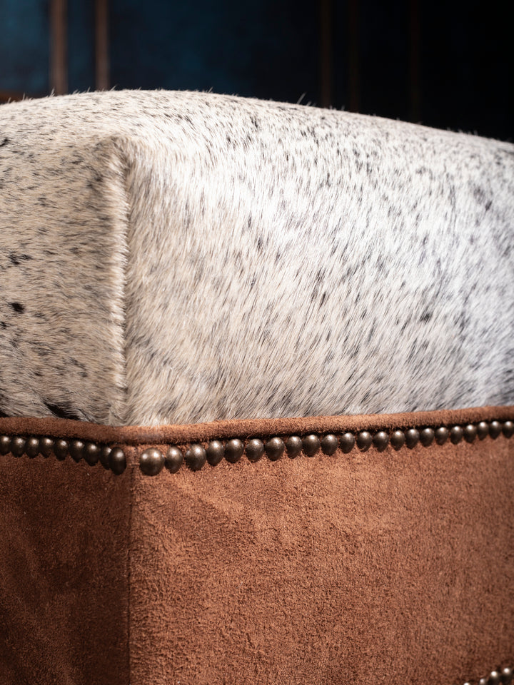 Angled view of square cowhide ottoman highlighting cowhide top, brown suede sides, and nailhead trim