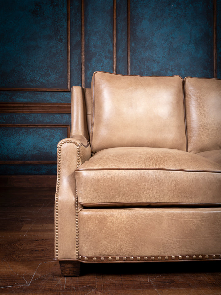 Close-up of the left side of the tan leather sofa, highlighting the armrest with nailhead trim and the plush cushion seat and backrest.