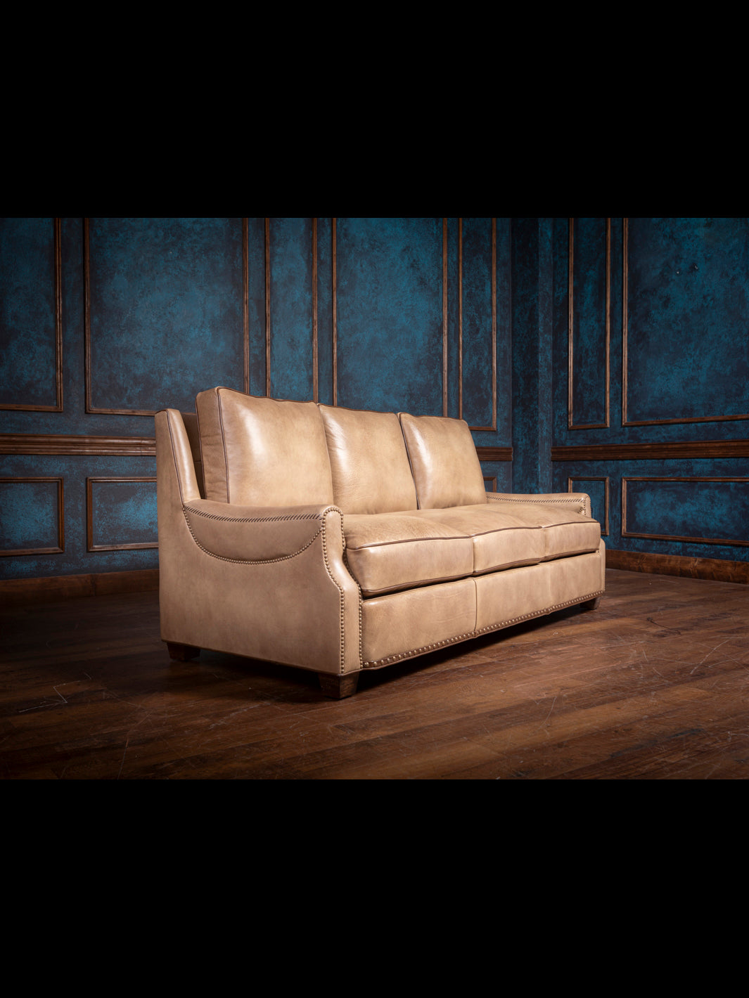 Side view of the tan leather sofa, showing smooth lines and sturdy construction, with a wooden floor and dark blue paneled wall backdrop.