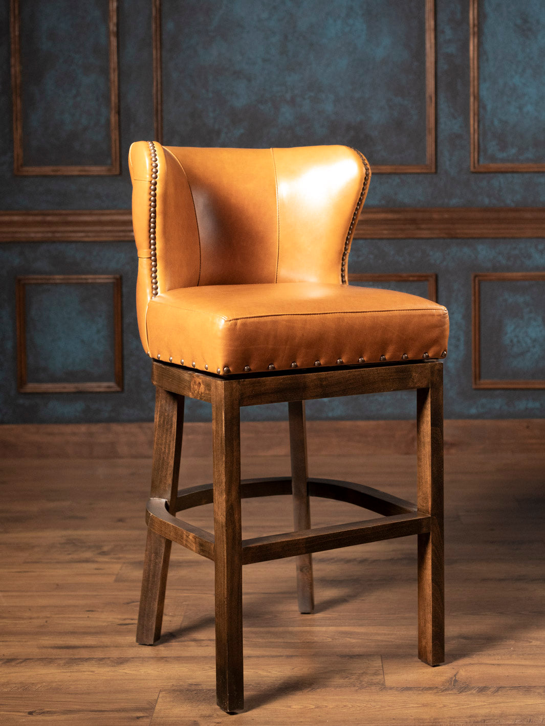 Brown leather bar stool with wooden legs against a textured wall.
