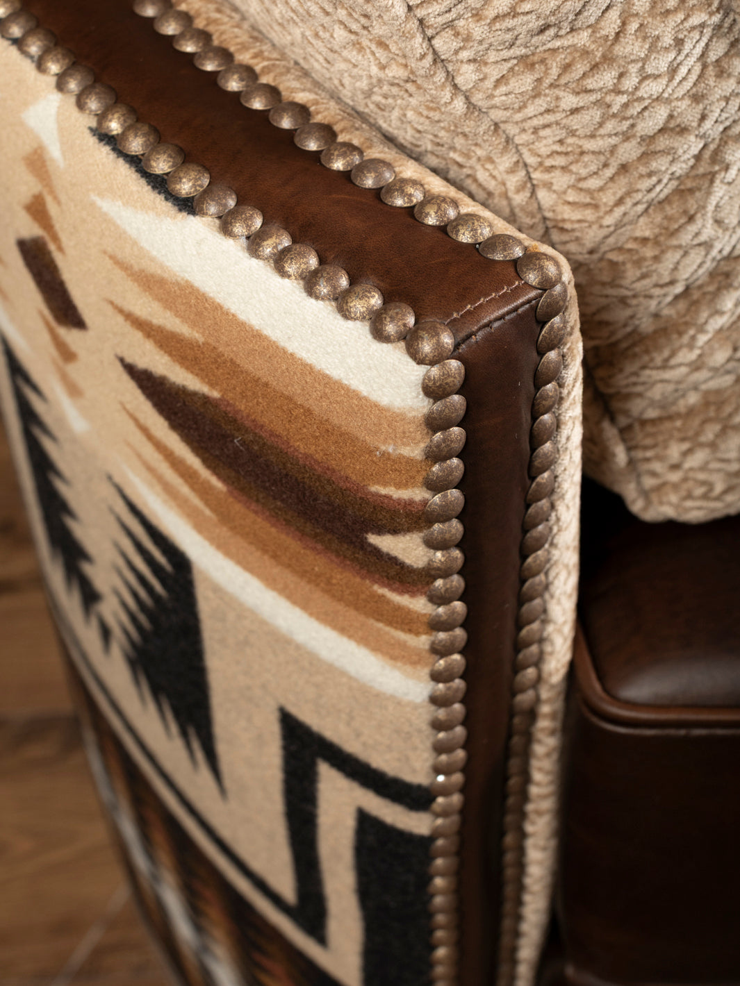 Detailed close-up shot of the single-track antiqued bronze nailhead stud trim running along the hand-burnished brown leather border of the southwestern patterned chair arm.