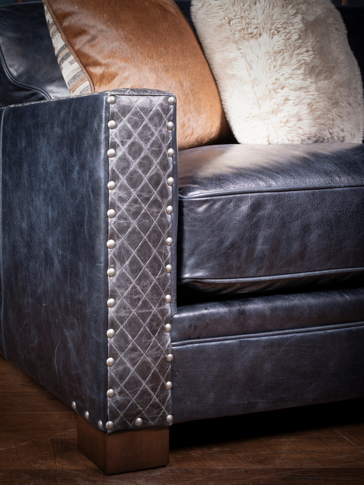 A close-up view of a luxurious blue leather sofa arm, showcasing intricate diamond-patterned quilted leather with large metallic nailhead trim. The leather has a rich, deep hue with a slightly distressed finish, adding to its vintage, western appeal. The sofa is complemented by accent pillows in earthy tones, adding texture and warmth to the overall aesthetic.