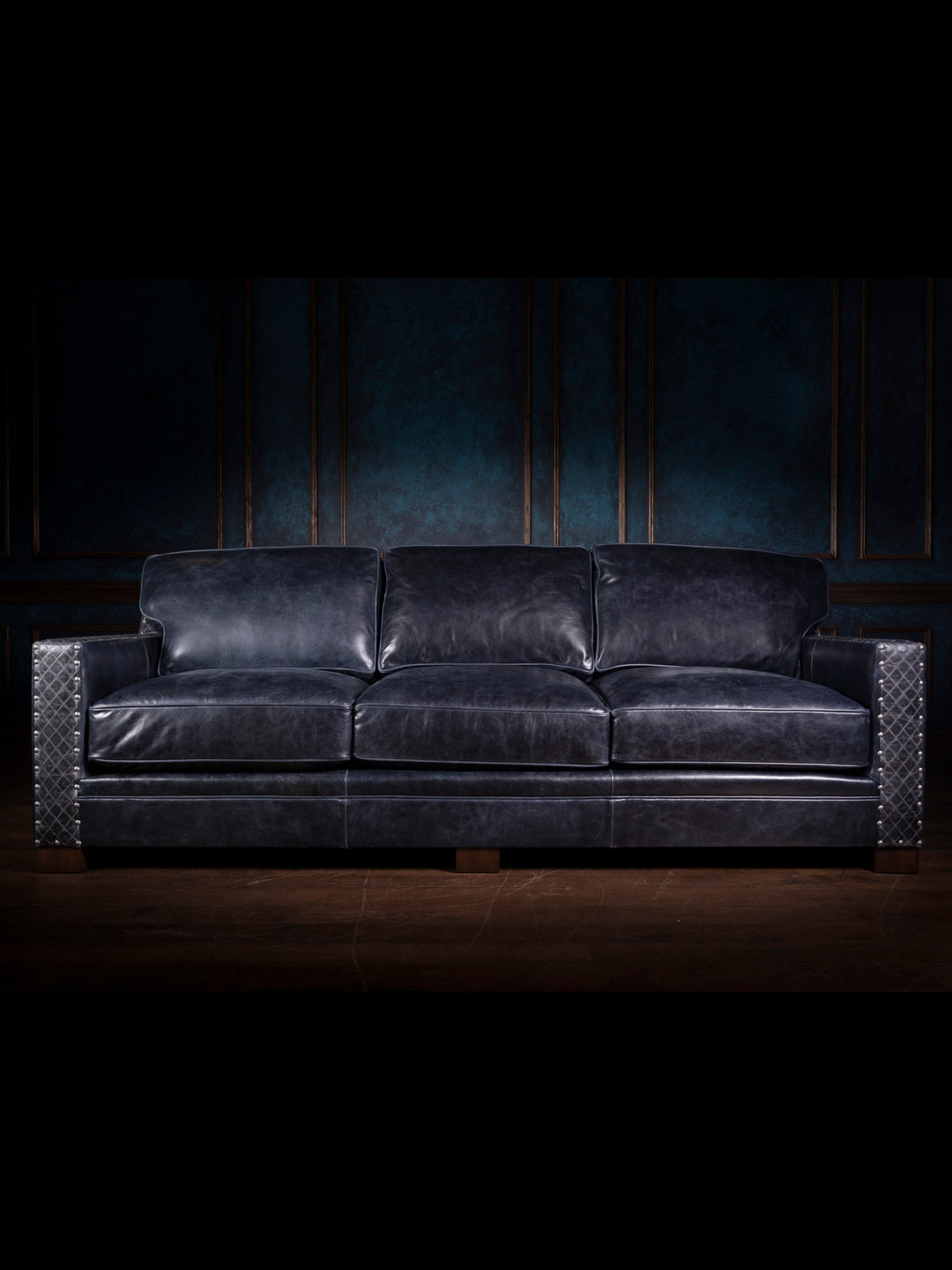 A front-facing shot of a three-seater blue leather sofa. The sofa's clean lines are softened by the plushness of the cushions and the rich, deep blue color of the leather. The arms of the sofa feature quilted leather panels with metallic nailhead trim, giving it a distinctive western flair. The sofa is set against a dark, textured wall, creating a dramatic contrast that highlights its luxurious design.