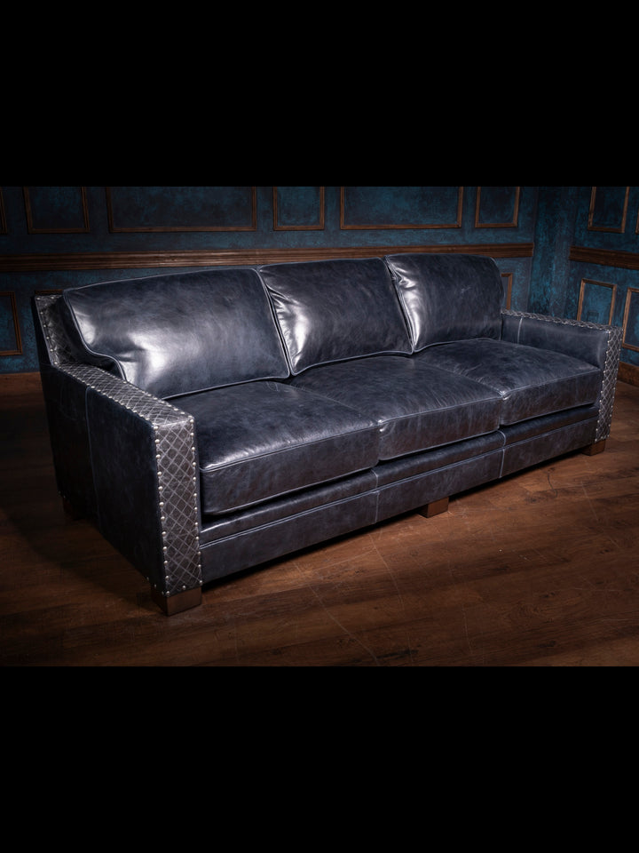 A side angle view of a blue leather sofa with quilted leather detailing on the arms. The sofa has a sleek design, with plush cushions that provide both comfort and style. The quilted panels on the arms are accentuated by large metallic nailhead trim, adding a touch of rugged sophistication. The deep blue leather has a slightly worn, vintage look, perfect for a rustic or western-themed interior.