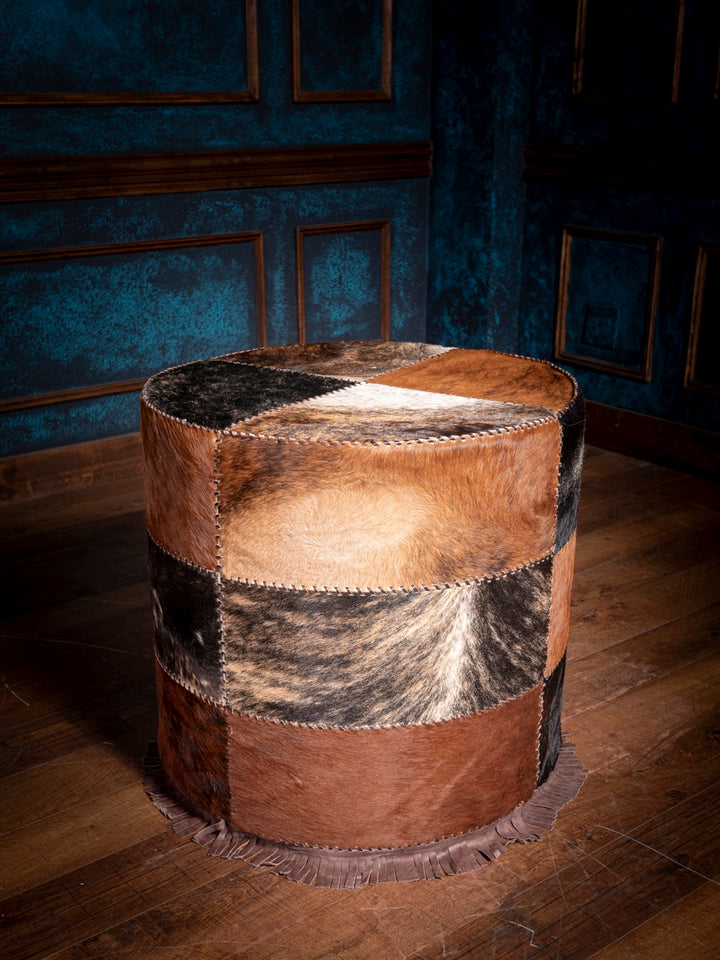 Round leather ottoman with patchwork design on a wooden floor against a blue paneled wall.