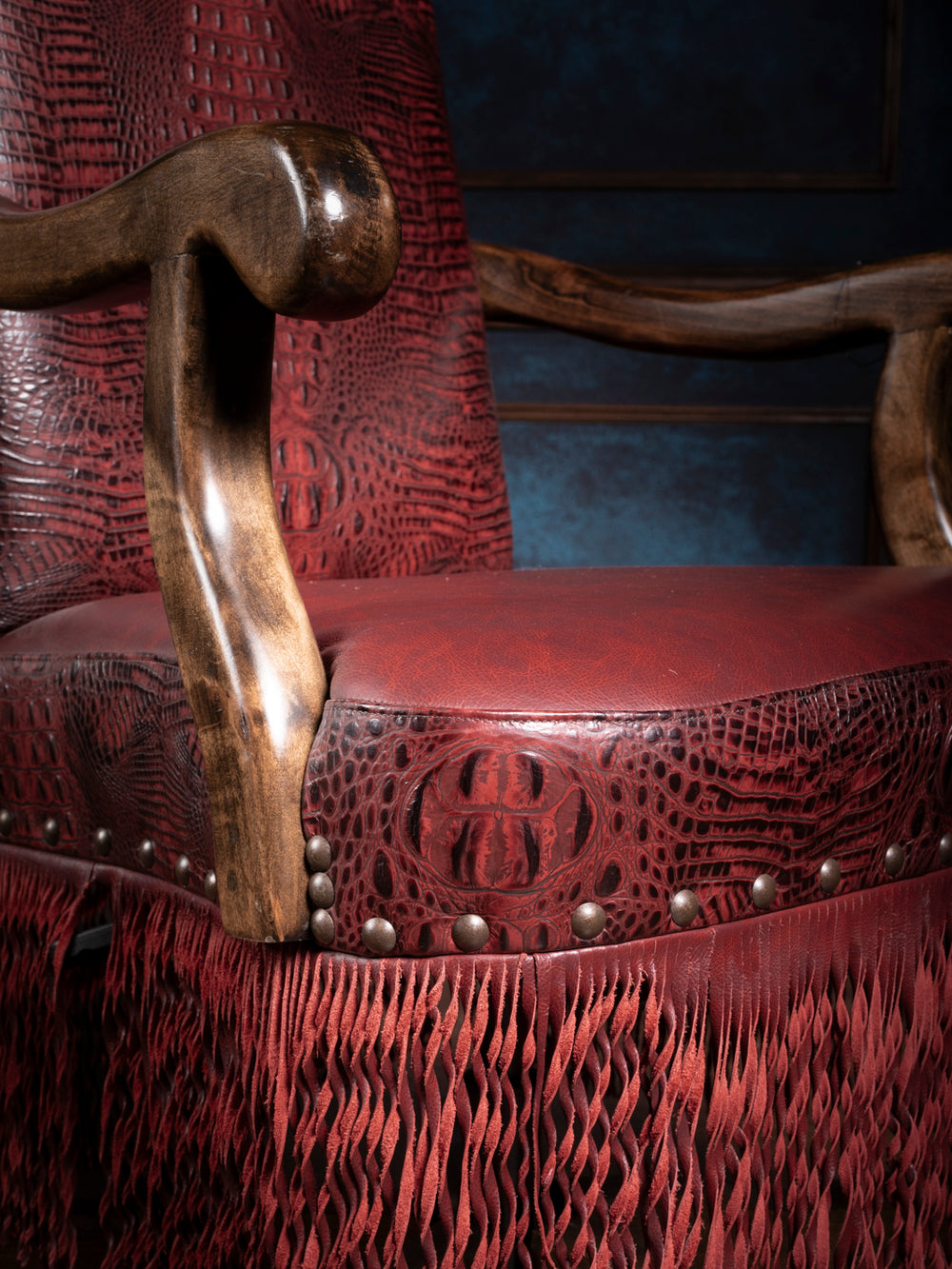 Close-up of crimson fringe desk chair detailing embossed leather, brass nailheads, and curved wooden armrest.