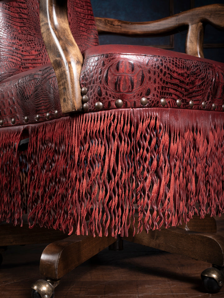 Angled view of a high-end crimson leather desk chair with twisted leather fringe, emphasizing the embossed pattern and brass nailhead accents.