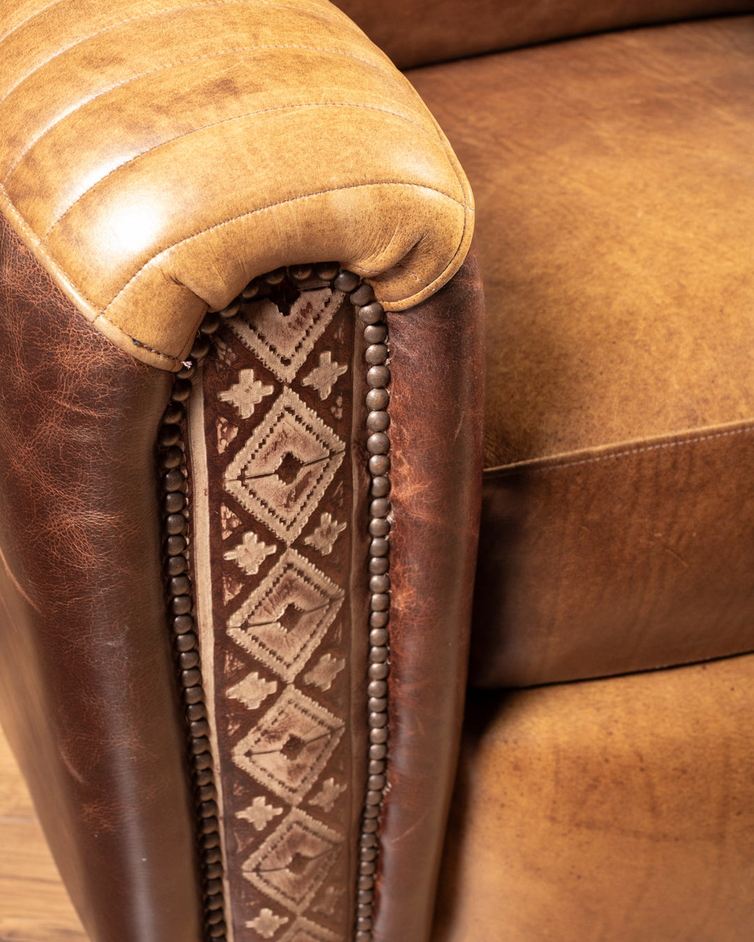 Macro detail of the intricate diamond hand-tooled geometric leather track accents and antique bronze nailhead trim on the arm facing.
