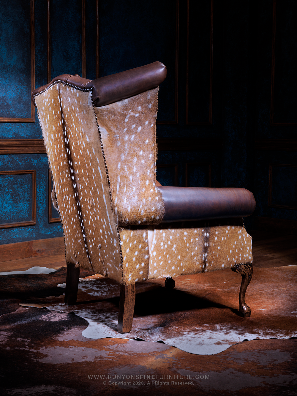 Elegant Axis Leather Accent Chair in Red Oak full-grain leather with Axis hair-on-hide back panels, wingback silhouette, and carved wood legs, shown from the rear in a rustic living room setting.