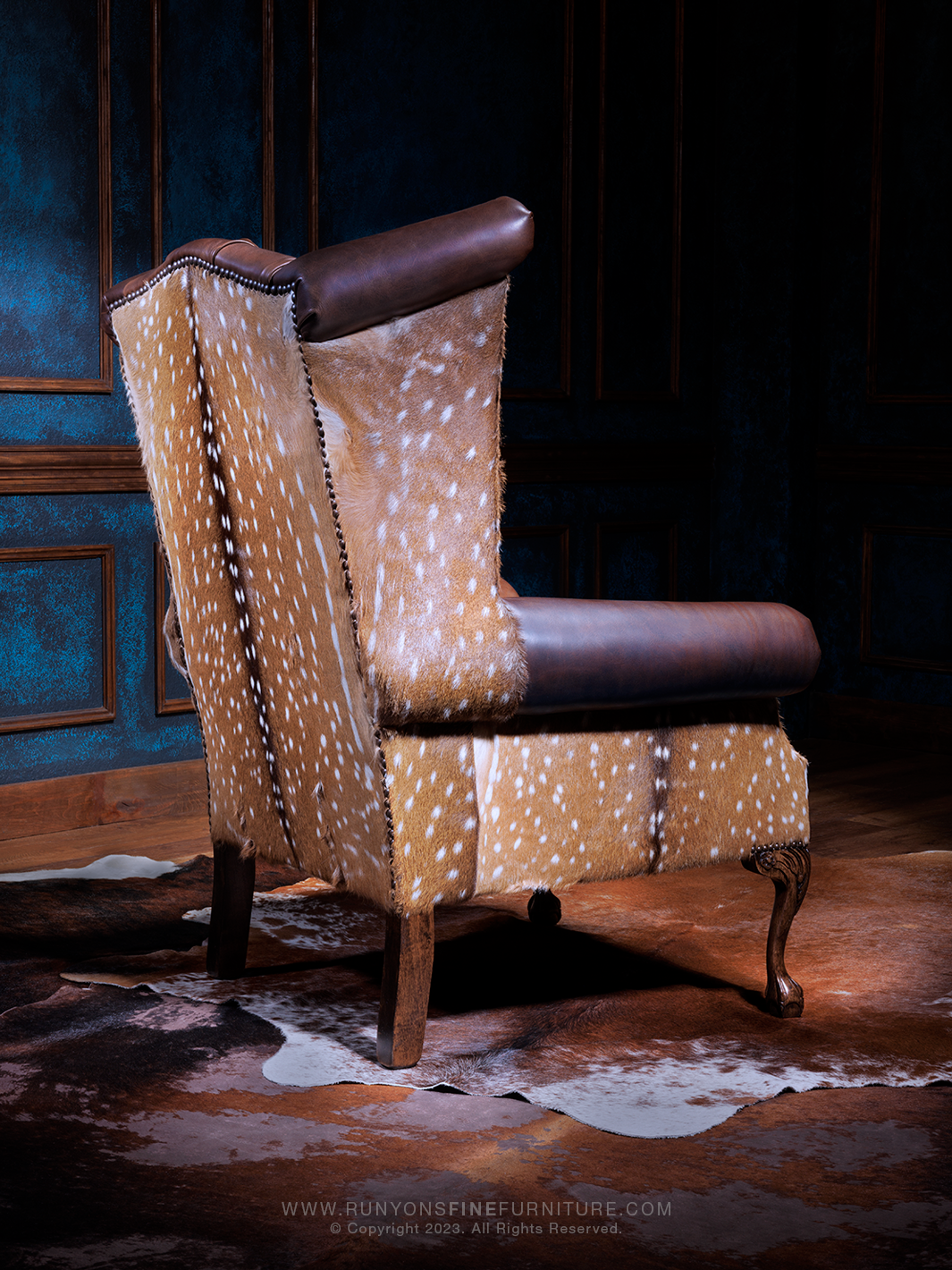Elegant Axis Leather Accent Chair in Red Oak full-grain leather with Axis hair-on-hide back panels, wingback silhouette, and carved wood legs, shown from the rear in a rustic living room setting.