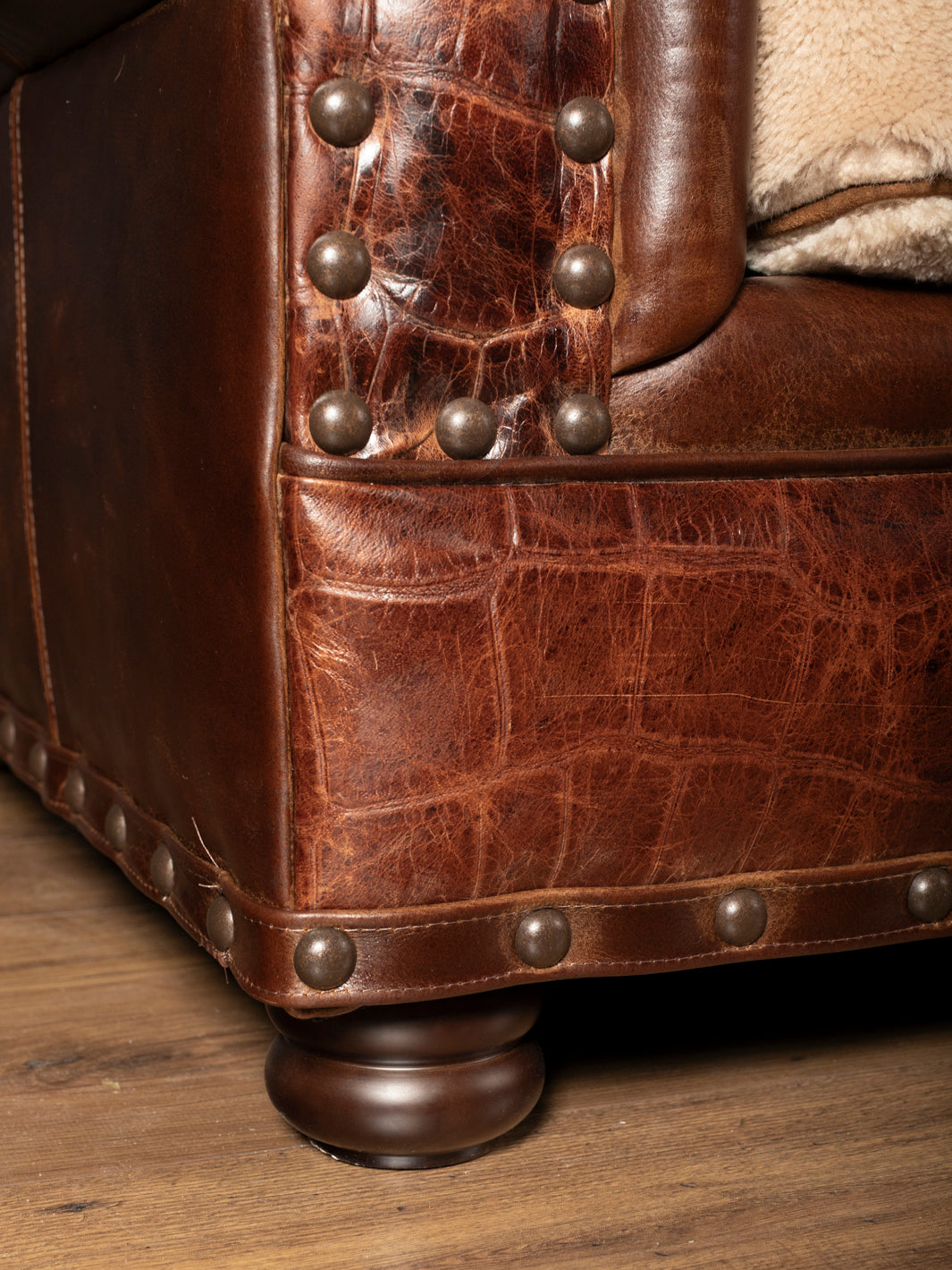 Close-up of a rustic sofa base featuring crocodile embossed leather panels, dark bronze nailhead trim, and a turned wooden foot.