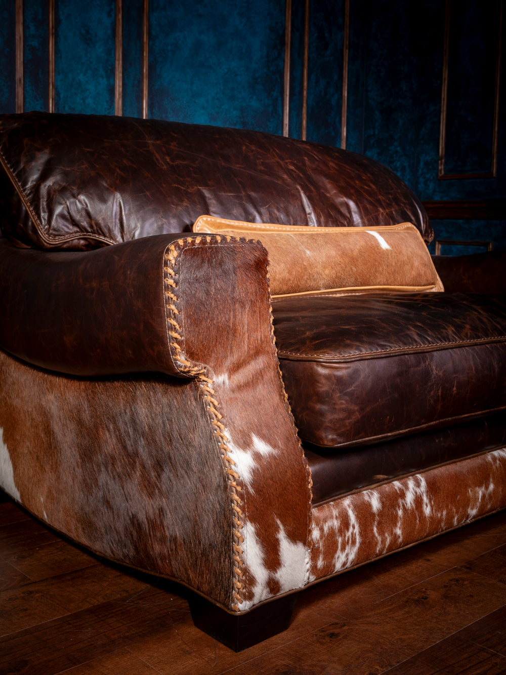 Close-up of a Western cowhide and leather chair-and-a-half, featuring distressed chocolate leather cushions, hair-on-hide sides, and cross-stitched whipstitch detailing.