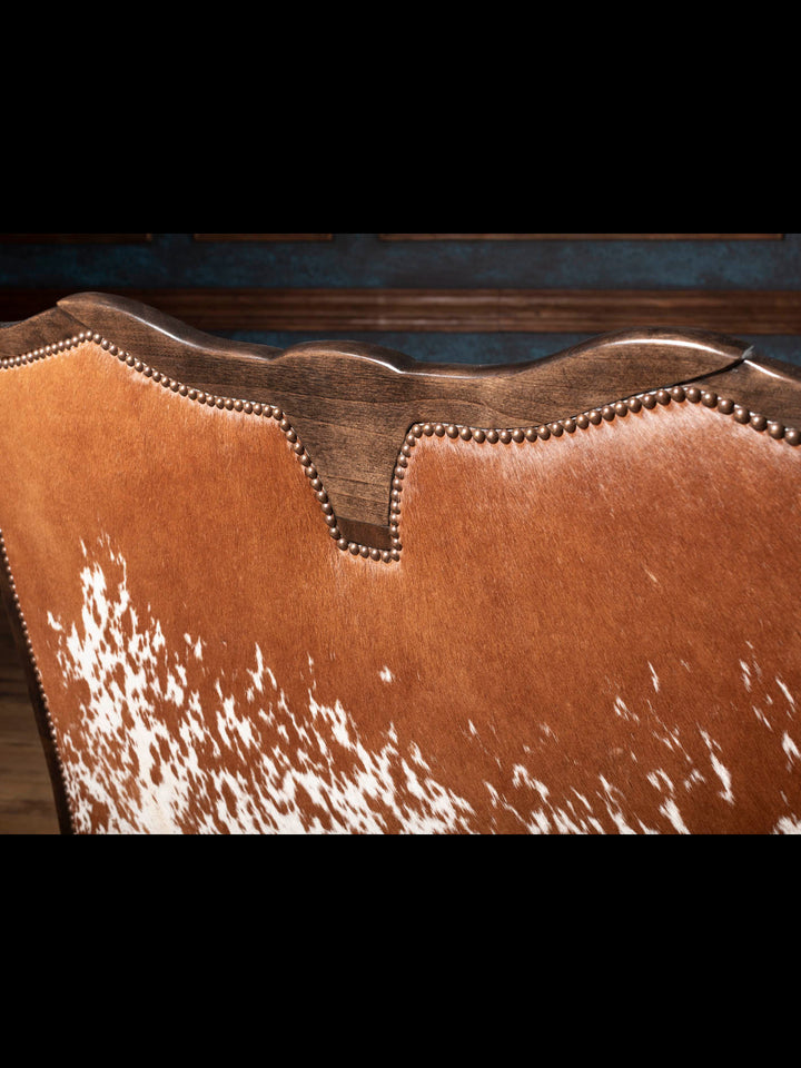 Close-up of the Longhorn Leather Bench featuring a genuine speckled red hair-on-hide back, hand-carved hardwood crest, and decorative brass nailhead trim.