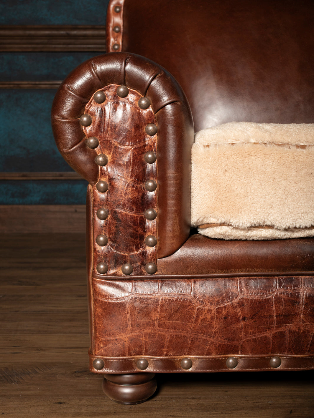 Detailed view of a brown leather rolled arm sofa panel with crocodile texture trim, brass nailheads, and shearling cushion.