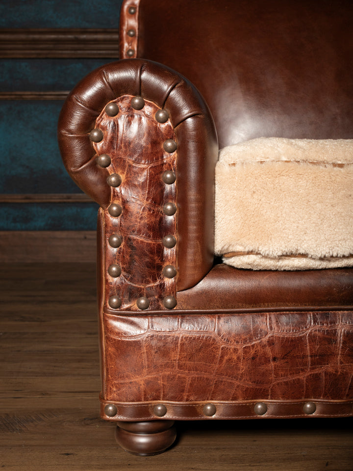 Detailed view of a brown leather rolled arm sofa panel with crocodile texture trim, brass nailheads, and shearling cushion.