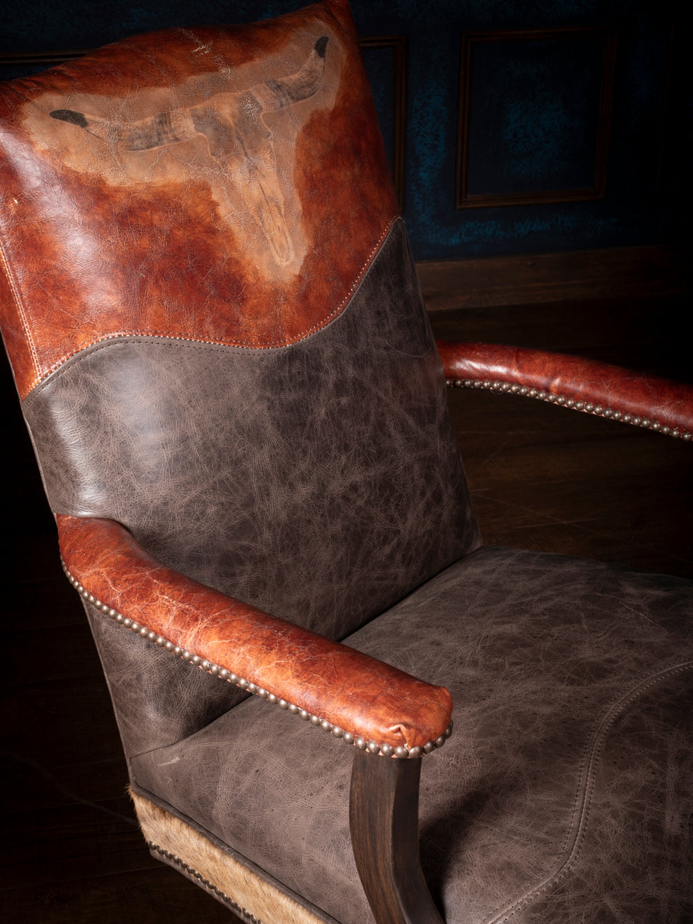 Close-up of a longhorn leather desk chair featuring a hand-tooled longhorn design on the backrest, distressed leather upholstery, and nailhead trim on the arms.