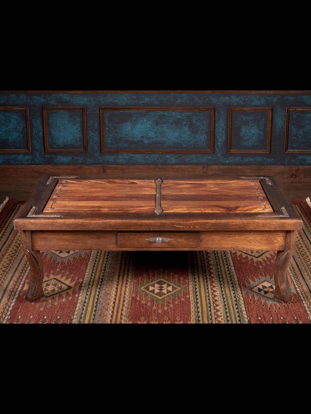 Wooden coffee table with a iron handle on a patterned rug against a dark wall.