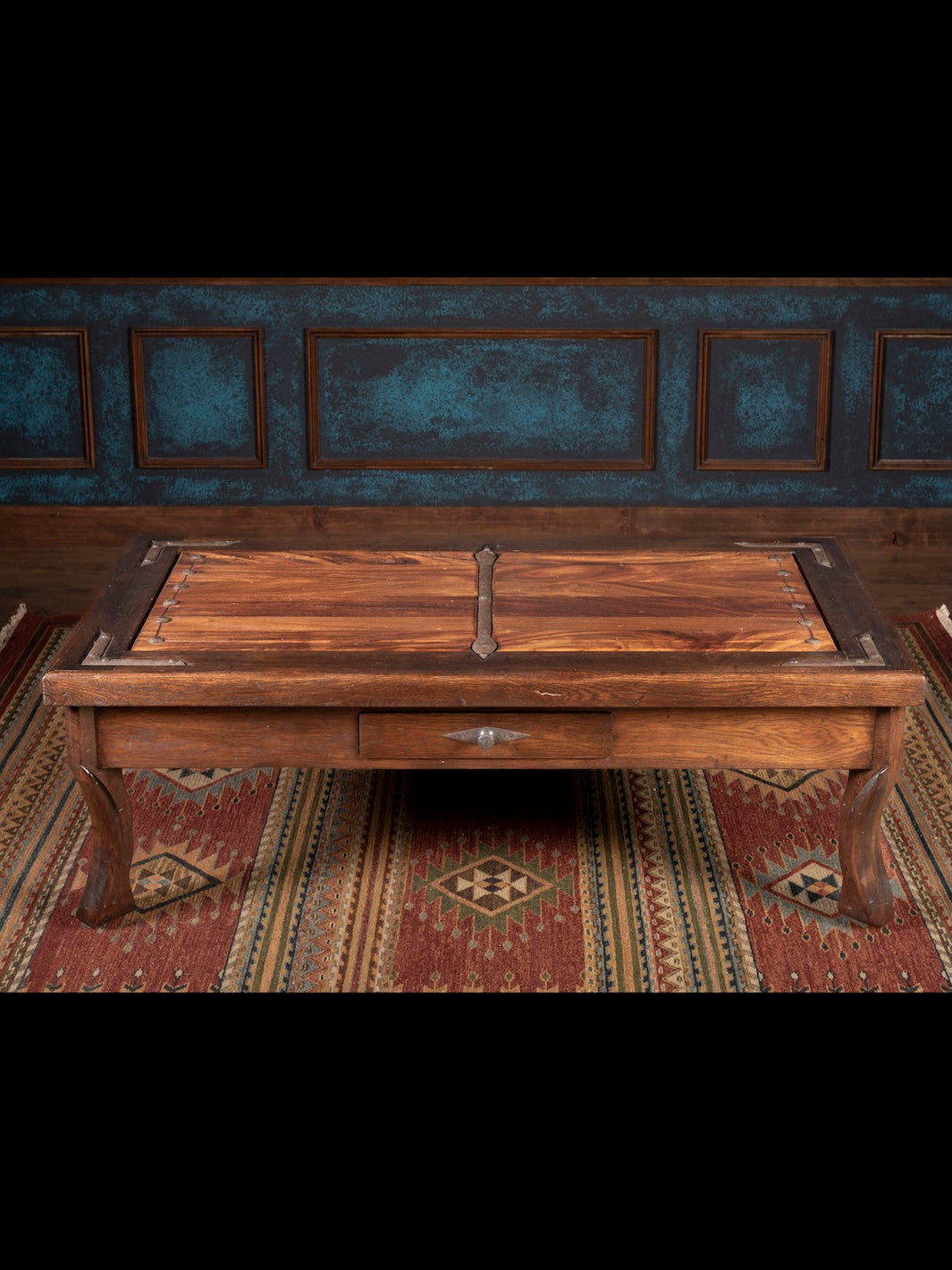 Wooden coffee table with a iron handle on a patterned rug against a dark wall.