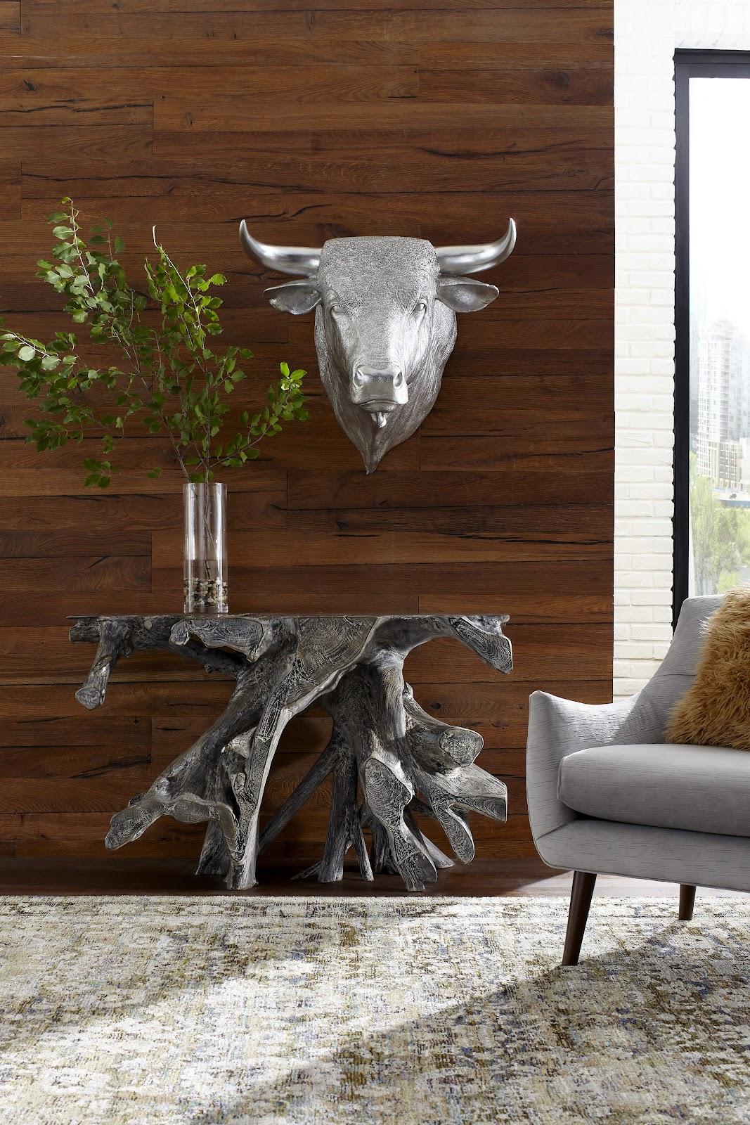 Decorative bull head on a wooden wall above a rustic console table with a vase and plant, next to a gray sofa.