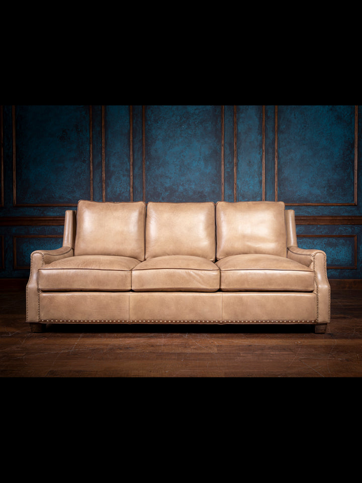 Tan leather three-seater sofa with nailhead trim detailing, set against a dark blue and wood-paneled wall. The sofa has plush cushions and a classic design.