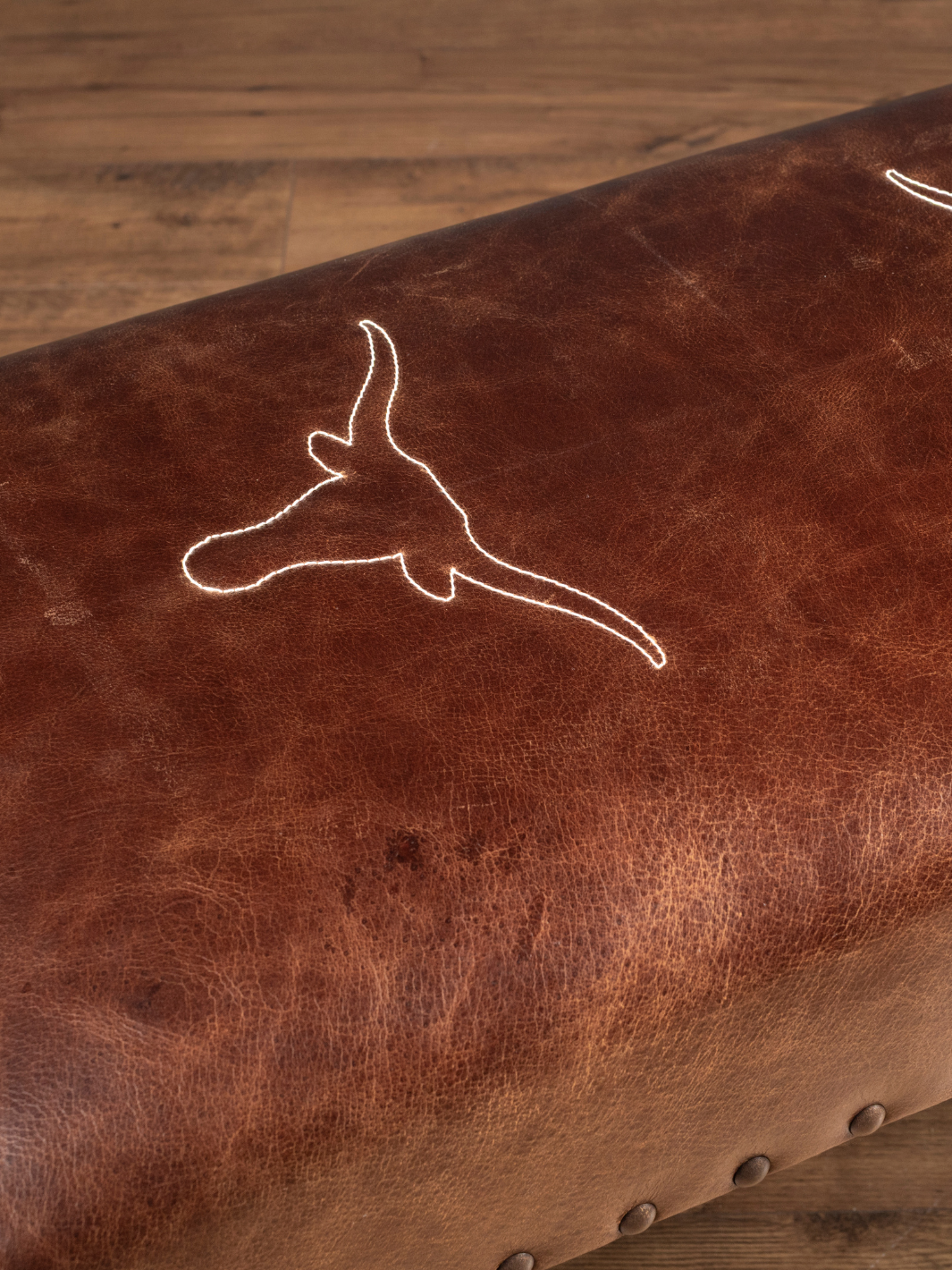 Close up view of the stitched Texas longhorn embroidery emblem on a full-grain buffalo leather bench seat, showcasing the natural hide grain and cream-colored artisan stitching.
