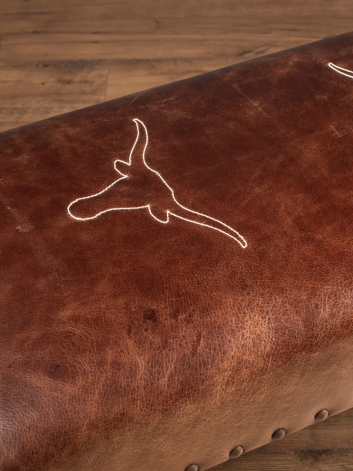 Close up view of the stitched Texas longhorn embroidery emblem on a full-grain buffalo leather bench seat, showcasing the natural hide grain and cream-colored artisan stitching.