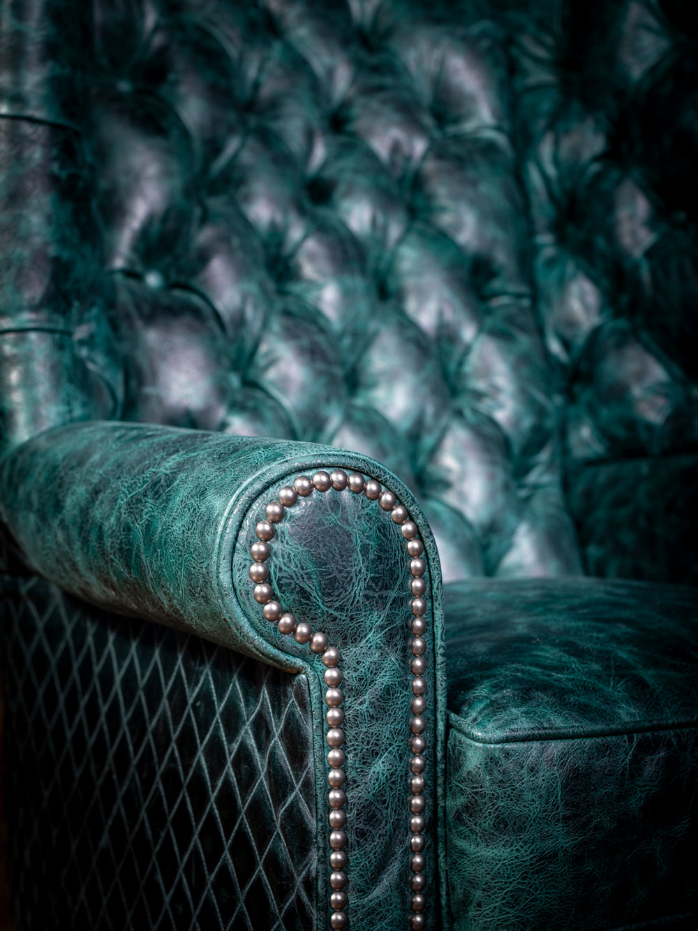 Tufted Teal Leather Wingback Chair in teal leather with diamond stitch accents featuring rolled arm design with silver nailheads in a close-up view.