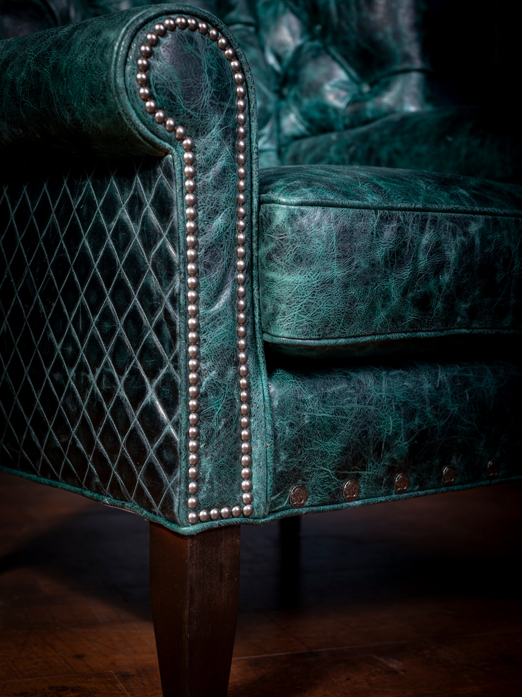 Tufted Leather Wingback Chair with full grain distressed teal leather and hand stitched diamond pattern featuring silver nailheads and dark brown straight feet in a close-up view.