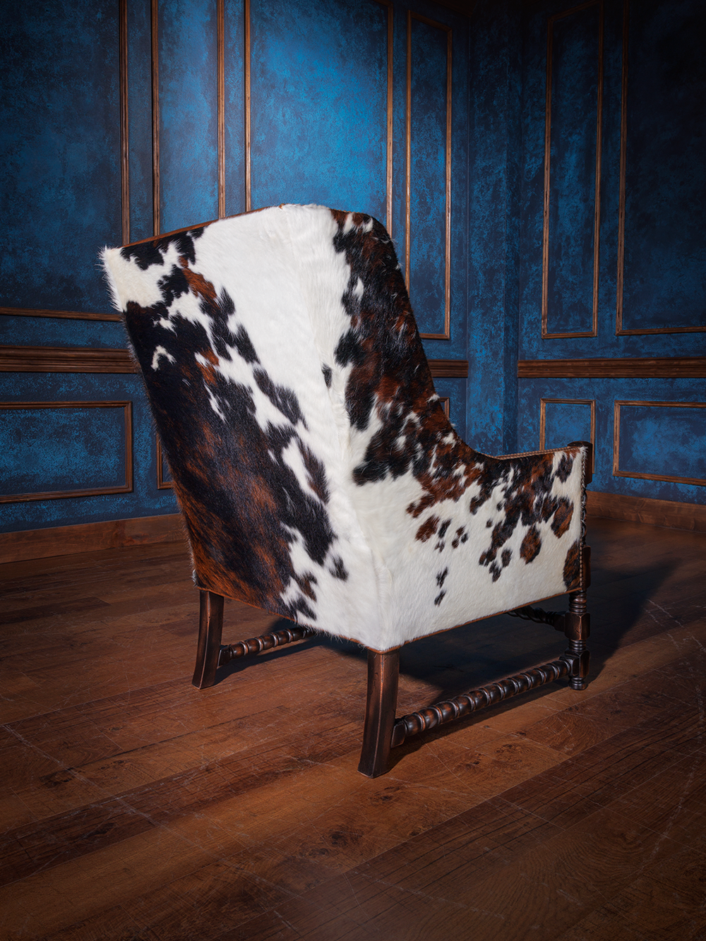 Rear view of the western-style armchair fully wrapped in tri-color cowhide, showing off its bold hide pattern and gently curved silhouette over a wooden floor.
