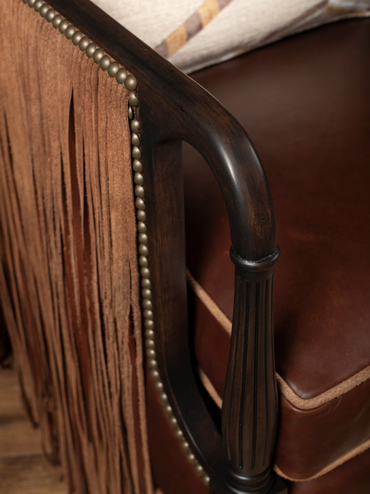 Close-up detail of hand-cut suede fringe curtains and traditional antique bronze nailhead trim tracking down a dark-washed wooden armrest frame.