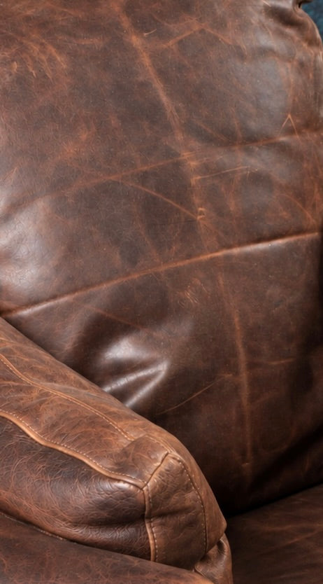 Close-up of a distressed brown leather chair back and arm showing surface scratching and creasing from daily contact