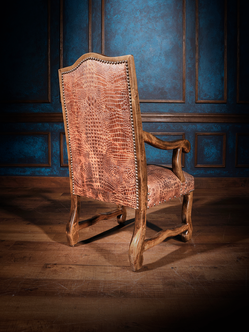 The back view of a wooden armchair featuring crocodile-embossed leather upholstery on both the seat and backrest, accented with nailhead trim, set against dark blue walls with wood paneling.