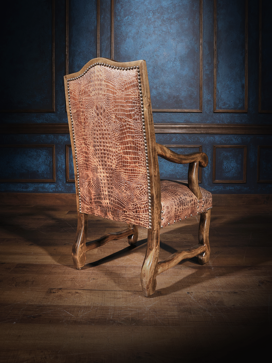 The back view of a wooden armchair featuring crocodile-embossed leather upholstery on both the seat and backrest, accented with nailhead trim, set against dark blue walls with wood paneling.