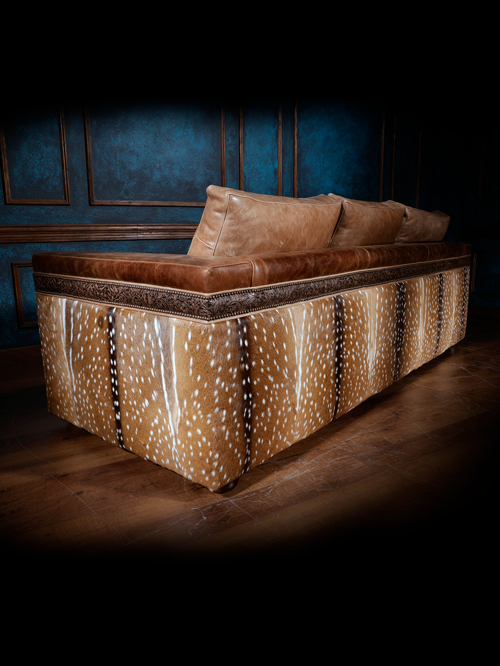 Back view of a western-style sofa with smooth tan leather cushions and intricate leather trim, featuring sides and back upholstered with axis deer hide, set against dark blue walls and wooden flooring.