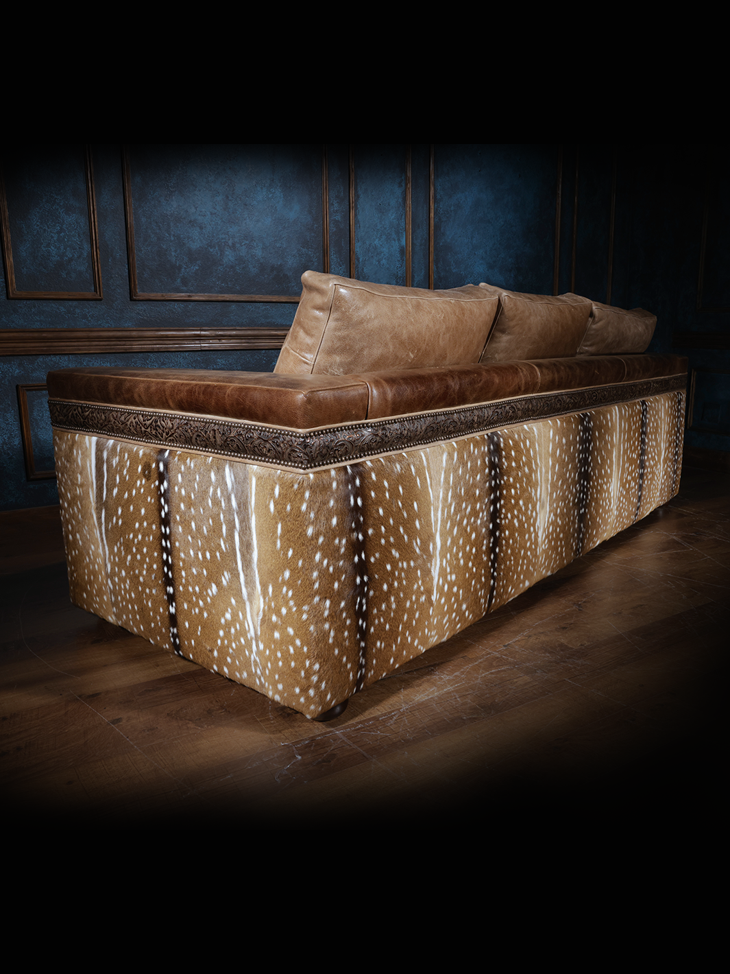 Back view of a western-style sofa with smooth tan leather cushions and intricate leather trim, featuring sides and back upholstered with axis deer hide, set against dark blue walls and wooden flooring.