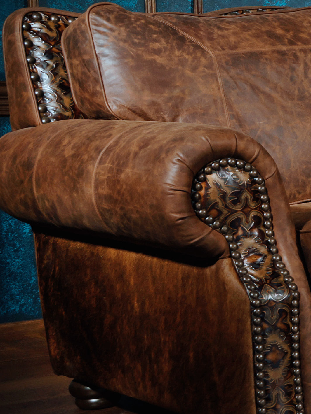 Close-up of a western-style sectional sofa armrest, showcasing intricate leather detailing with nailhead trim and brown leather upholstery.