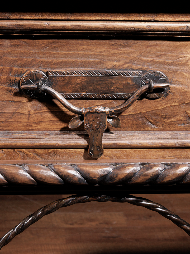 Detail of a drawer handle shaped like a longhorn, on a rustic wooden console table, highlighting the intricate metalwork and wood texture.