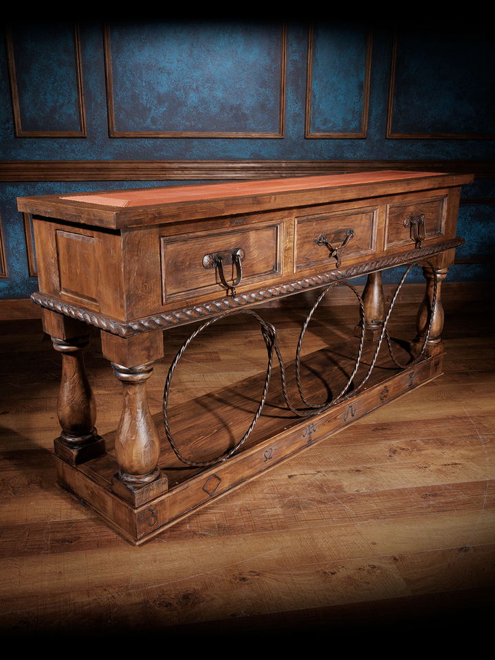 Rustic wooden console table with a brown leather inlay, metal ring accents, and ornate carvings, set against a dark wall background.