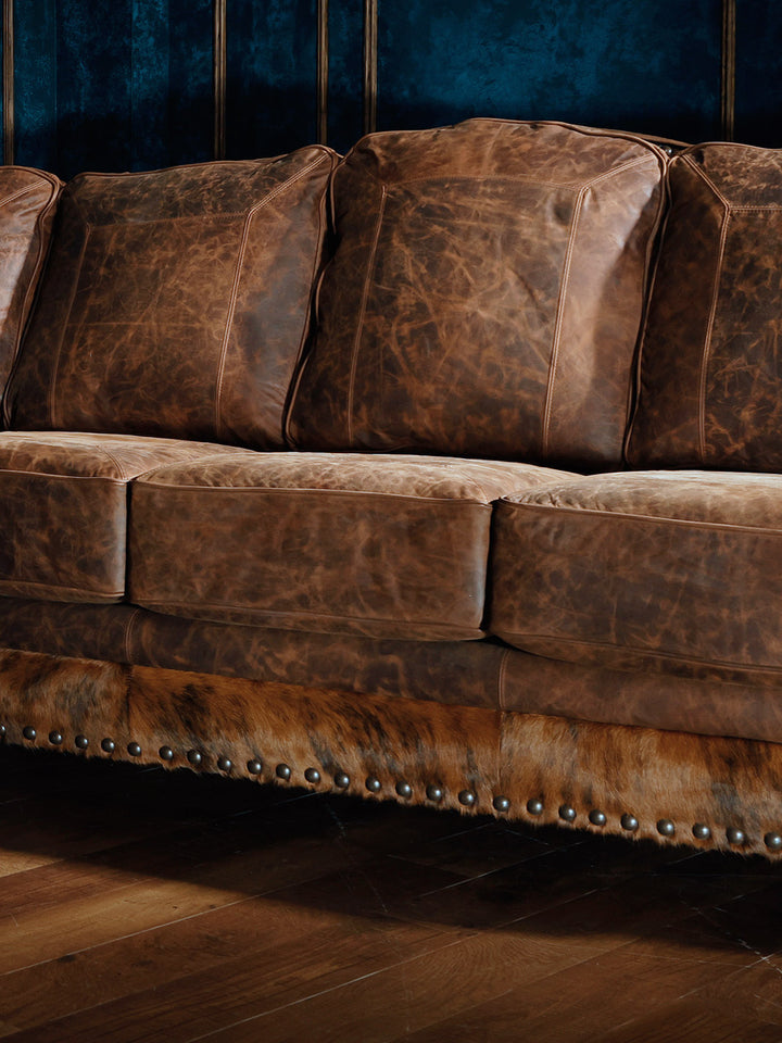Close-up of the seat cushions on a western-style sectional sofa, featuring distressed brown leather upholstery and hair-on-hide accents with nailhead trim along the base.