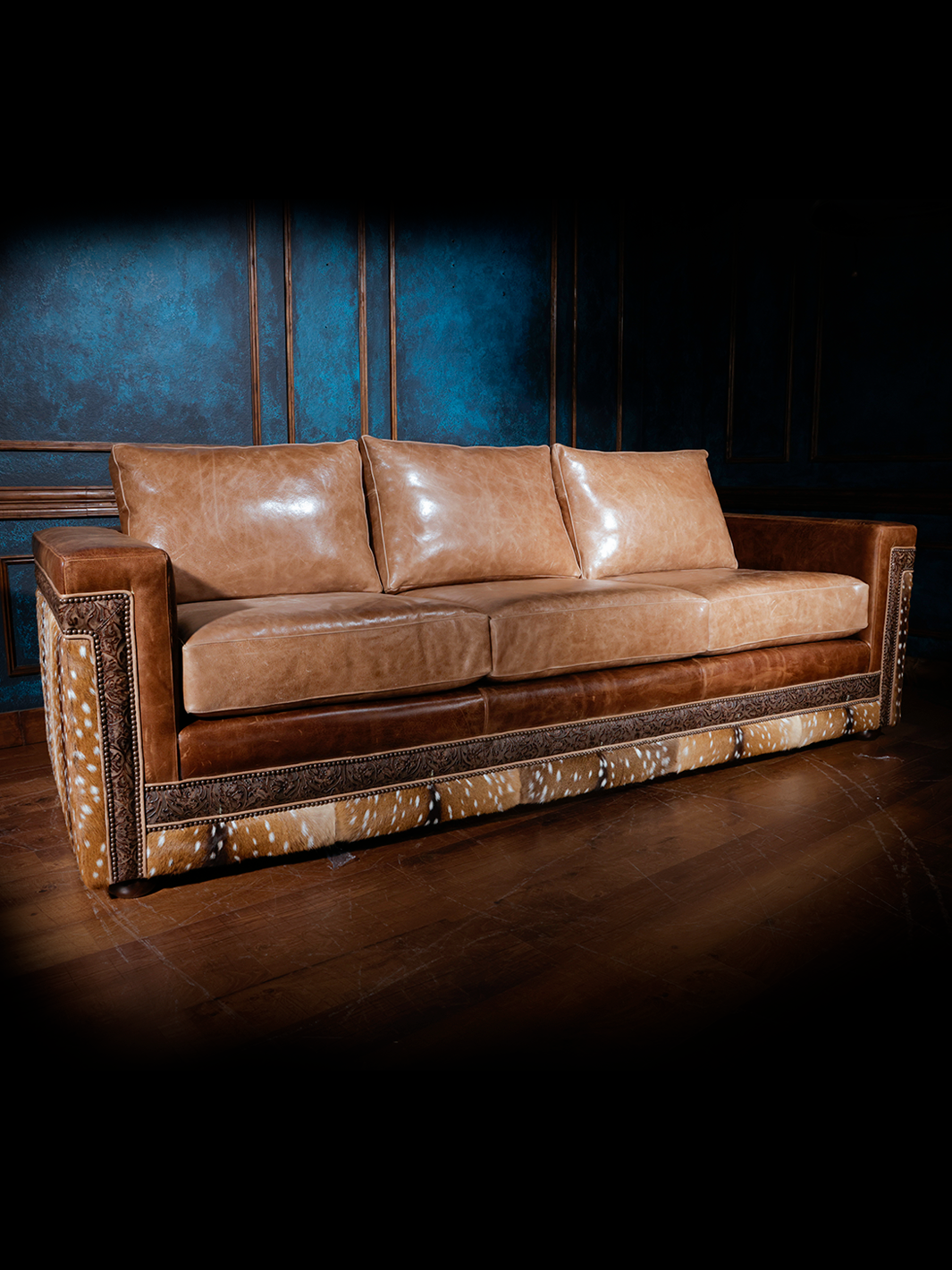 A western-style sofa featuring smooth tan leather cushions, detailed leather trim, and sides and back upholstered with axis deer hide, set in a room with dark blue walls and wooden flooring.