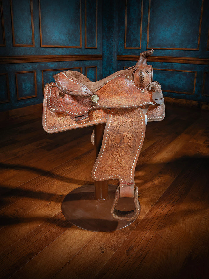 A detailed western-style saddle bar stool featuring intricate leather tooling and silver accents, mounted on a sturdy metal base designed to mimic the look and feel of a real saddle.